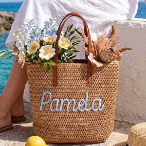 May include: A tan woven straw tote bag featuring the name Pamela embroidered in light blue script on the front. The bag has two brown leather handles and is filled with a variety of colourful flowers, including blue delphiniums and white roses.