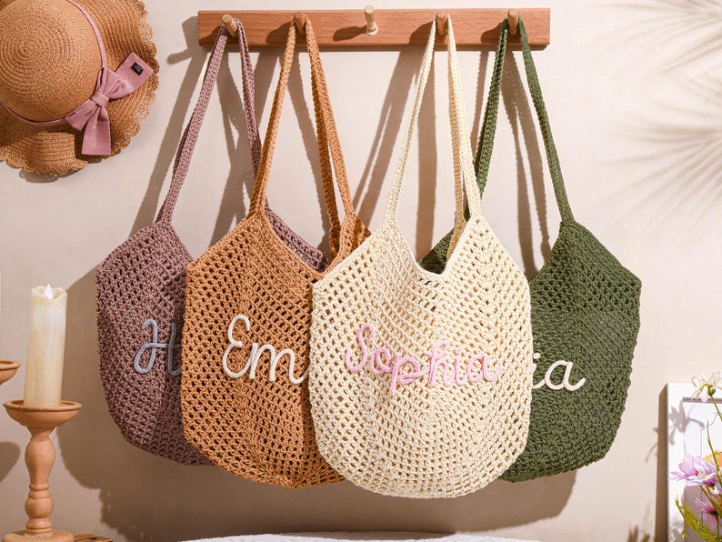 May include: Four crocheted tote bags in shades of purple, brown, cream, and green hang from a wooden rack. Each bag has a personalized name embroidered on the front in a contrasting color. A straw hat with a pink bow is visible in the background.