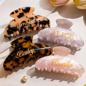 May include: Four personalised hair claw clips in tortoiseshell, lavender, and pink. Each clip has a name in gold lettering: Amelia, Bailey, Alice, and Rosalie. The clips are on a white surface with pearls and decorative items.