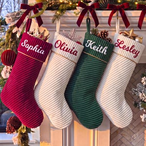 Könnte beinhalten: Vier gerippte Weihnachtsstrümpfe hängen in einer Reihe. Der erste ist dunkelrot mit dem Namen Sophia, der zweite weiß mit Olivia, der dritte dunkelgrün mit Keith und der vierte weiß mit Lesley. Jeder Strumpf hat eine Manschette und eine Samtschleife.