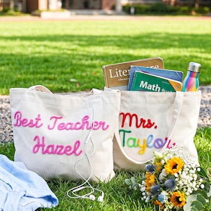 K&ouml;nnte beinhalten: Zwei cremefarbene Cord-Einkaufstaschen mit gesticktem Text. Eine Tasche zeigt "Best Teacher" und "Hazel" in rosa Garn. Die andere Tasche zeigt "Mrs." in Rot und "Taylor" in mehrfarbigem Garn und enth&auml;lt B&uuml;cher sowie eine regenbogenfarbene Trinkflasche.