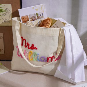 K&ouml;nnte beinhalten: Eine cremefarbene Cord-Einkaufstasche mit dem gestickten Text Mrs. Williams in rotem, rosa, gelbem, gr&uuml;nem, blauem und violettem Garn. Die Tasche enth&auml;lt eine Holzb&uuml;rste, einen wei&szlig;en Stoffartikel und mehrere Karten mit aufgedrucktem Text.