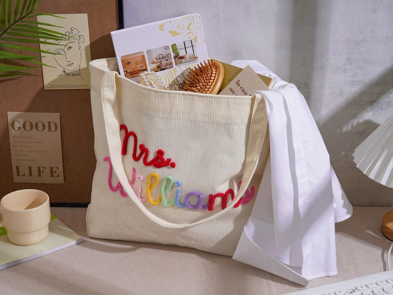 K&ouml;nnte beinhalten: Eine cremefarbene Cord-Einkaufstasche mit dem gestickten Text Mrs. Williams in rotem, rosa, gelbem, gr&uuml;nem, blauem und violettem Garn. Die Tasche enth&auml;lt eine Holzb&uuml;rste, einen wei&szlig;en Stoffartikel und mehrere Karten mit aufgedrucktem Text.