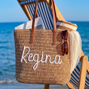 May include: A tan woven straw tote bag with brown leather handles and the name Regina embroidered in white script on the front. The bag contains a white book, a brown and white striped towel, and a pair of brown sunglasses hanging on the side.