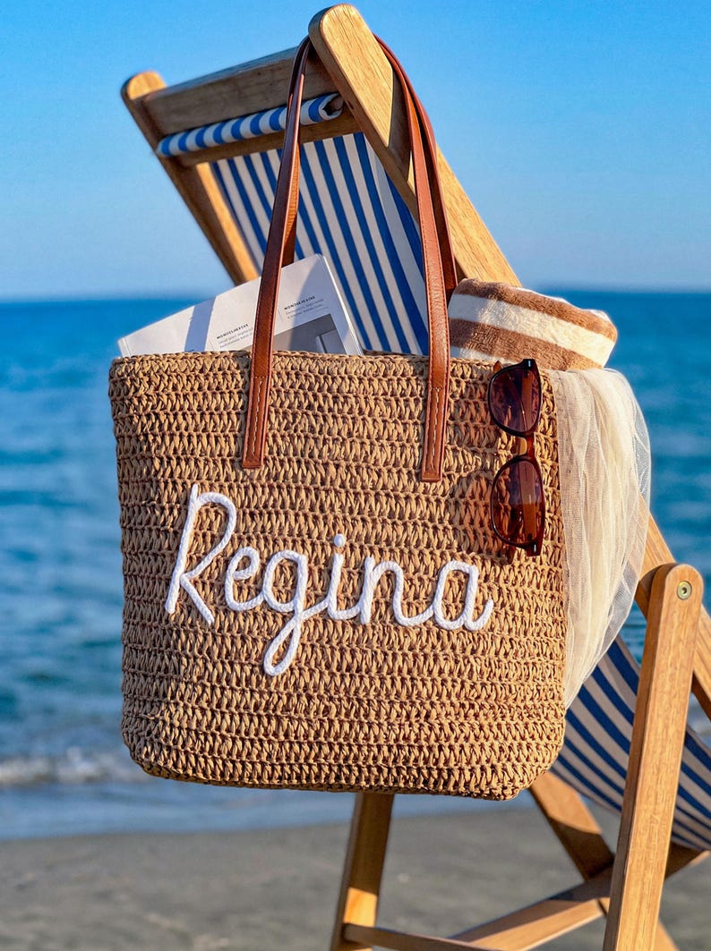 May include: A tan woven straw tote bag with brown leather handles and the name Regina embroidered in white script on the front. The bag contains a white book, a brown and white striped towel, and a pair of brown sunglasses hanging on the side.