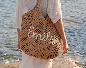 May include: A woven, tan tote bag with the name "Emily" embroidered in white script. The bag has a large, open weave design and is carried by a person wearing a white dress. The background shows the ocean.