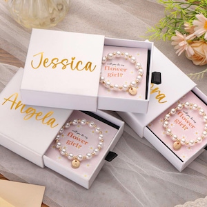 May include: White gift boxes with gold lettering, each containing a pearl bracelet with a gold charm. The boxes are labeled with names like "Jessica" and "Angela". The bracelets are presented on pink cards with the text "will you be my flower girl?"