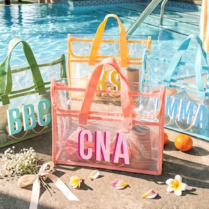 Peut inclure: Quatre sacs fourre-tout rectangulaires en plastique transparent avec des bordures et des poignées colorées. Chaque sac présente de grandes lettres monogrammes en bloc contrastées sur le devant. Les sacs sont disposés en groupe, avec des tons vert, jaune, corail et bleu.