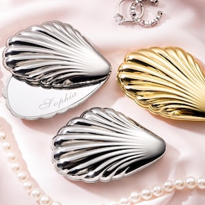 May include: Three seashell-shaped compact mirrors in silver and gold, arranged on a pink satin surface. One silver compact is open, revealing a mirror with the name "Sophia" engraved. A pearl necklace and a pair of earrings are also present.