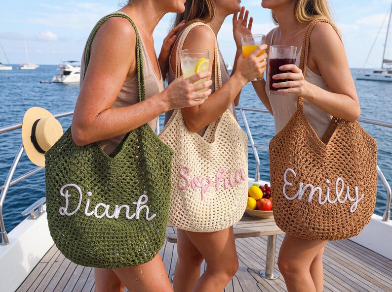 May include: Three open-weave tote bags in green, beige, and brown, each with a name embroidered in a contrasting color. The bags are held by people on a boat, with drinks and a hat visible. The names are Dianh, Sophia, and Emily.