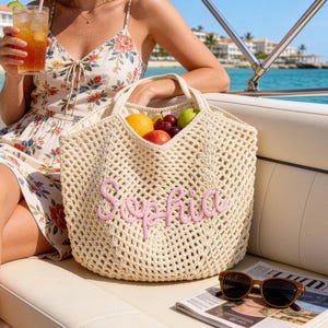 May include: A cream-colored, open-weave tote bag with the name "Sophia" embroidered in pink. The bag is filled with fresh fruit. Sunglasses and a newspaper rest on a white surface. The scene is set on a boat with a view of the ocean.