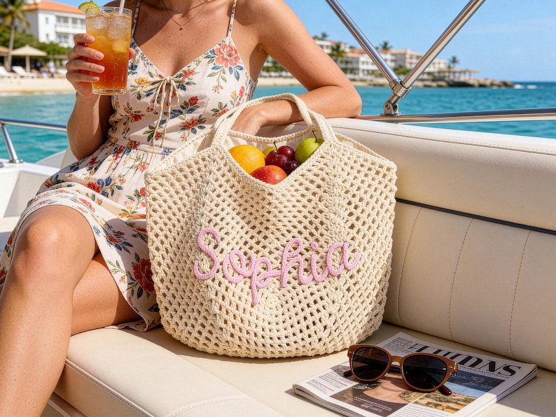 May include: A cream-colored, open-weave tote bag with the name "Sophia" embroidered in pink. The bag is filled with fresh fruit. Sunglasses and a newspaper rest on a white surface. The scene is set on a boat with a view of the ocean.