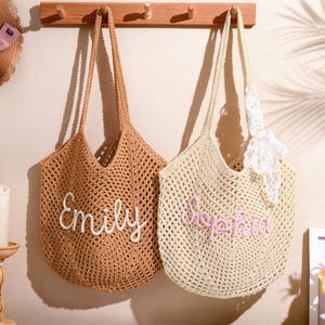 May include: Two crocheted tote bags, one tan and one cream, hang on a wooden rack. The tan bag has "Emily" embroidered in white, and the cream bag has "Sophia" in pink. A straw hat with a pink bow and candles are also visible.