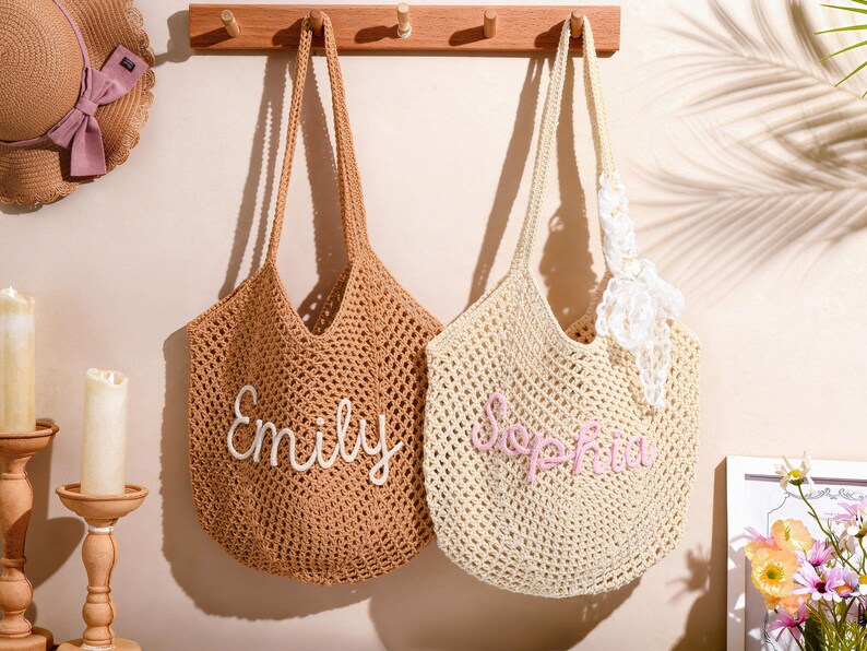 May include: Two crocheted tote bags, one tan and one cream, hang on a wooden rack. The tan bag has "Emily" embroidered in white, and the cream bag has "Sophia" in pink. A straw hat with a pink bow and candles are also visible.
