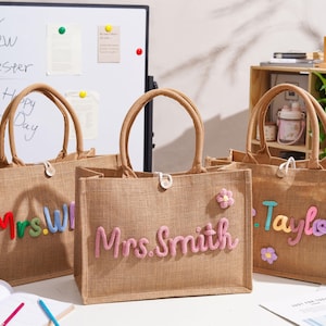 May include: Three rectangular jute tote bags with brown handles and a single button closure. Each bag features colorful, raised, fuzzy lettering displaying names like Mrs. Smith and Taylor, accented with small, soft, multi-colored flower patches.