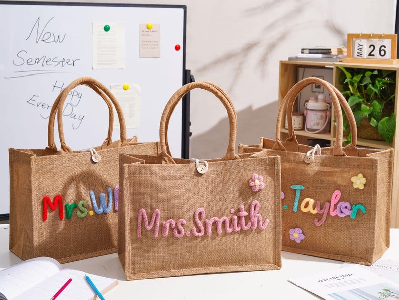 May include: Three rectangular jute tote bags with brown handles and a single button closure. Each bag features colorful, raised, fuzzy lettering displaying names like Mrs. Smith and Taylor, accented with small, soft, multi-colored flower patches.