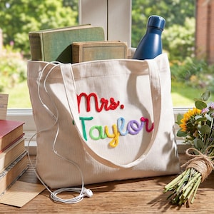 K&ouml;nnte beinhalten: Eine cremefarbene Cord-Tragetasche mit dem gestickten Text Mrs. Taylor in rotem, gr&uuml;nem, gelbem und violettem Garn. Die Tasche enth&auml;lt zwei Hardcover-B&uuml;cher und eine dunkelblaue Wasserflasche, davor liegen wei&szlig;e kabelgebundene Ohrh&ouml;rer.