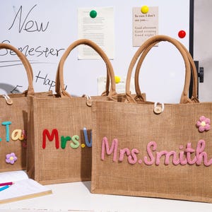 May include: Three rectangular jute tote bags with brown handles and a button closure. Each bag features colorful, raised, cursive-style lettering spelling out names like Mrs. Smith, along with small, decorative, multi-colored fabric flower appliques.