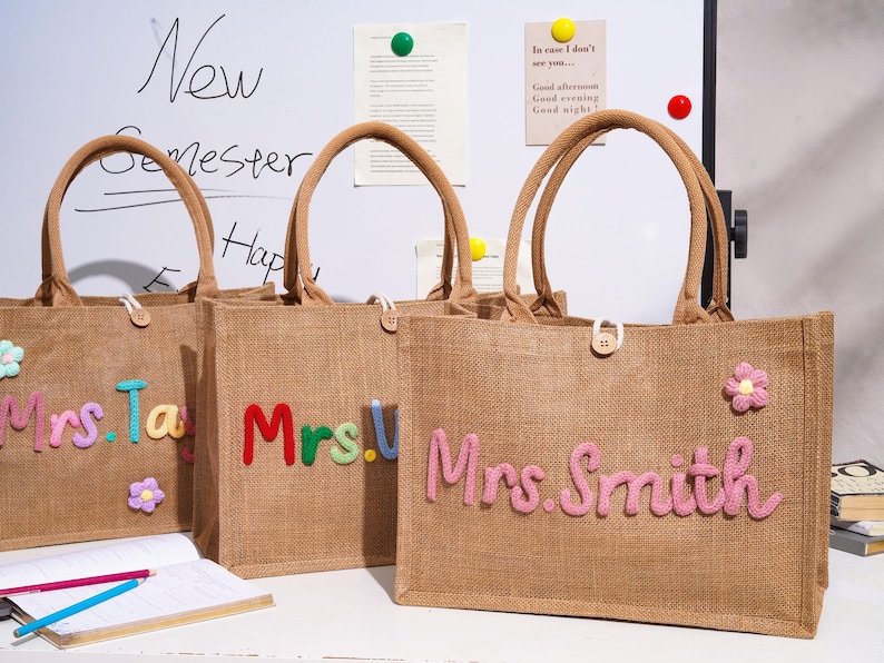 May include: Three rectangular jute tote bags with brown handles and a button closure. Each bag features colorful, raised, cursive-style lettering spelling out names like Mrs. Smith, along with small, decorative, multi-colored fabric flower appliques.