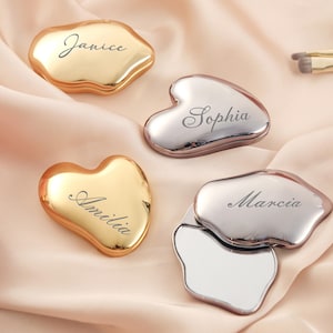 Op de afbeelding: Vier gepersonaliseerde metalen Gua Sha-tools in goud en zilver, elk gegraveerd met een naam: Janice, Amelia, Sophia en Marcia. De tools worden gepresenteerd op een zachte, perzikkleurige stof, met make-up kwasten op de achtergrond.