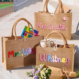 May include: Three rectangular tan jute tote bags with two top handles and a single wooden button closure. Each bag features colorful, raised, embroidered lettering on the front: one reads Mrs. Smith, one reads Mrs. Whit, and one reads Whitney.
