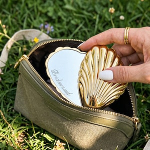 May include: A gold seashell-shaped compact mirror with the word "Bridesmaid" on the mirror's surface is held open next to a green canvas zippered pouch. The compact is gold and has a scalloped edge. The pouch is olive green.