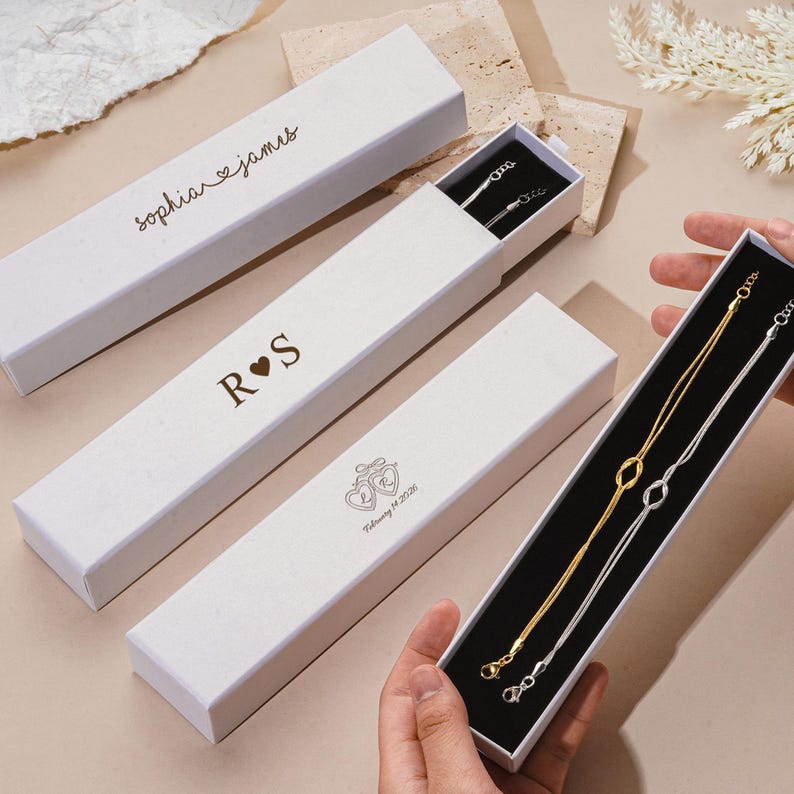 K&ouml;nnte beinhalten: Wei&szlig;e rechteckige Geschenkboxen mit individuellem Text und goldenen oder silbernen Kettenhalsketten. Eine Box tr&auml;gt die Aufschrift "sophia & james", eine andere "R &hearts; S", und die dritte ein Herzdesign und "February 14 2026".