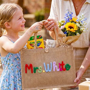 Könnte beinhalten: Eine Jute-Tragetasche mit dem Namen "Mrs. White" in bunten, strukturierten Buchstaben. Die Tasche ist mit Schulmaterialien und einer Tasse mit der Aufschrift "Best Teacher" gefüllt. Ein Blumenstrauß wird in der Nähe gehalten.