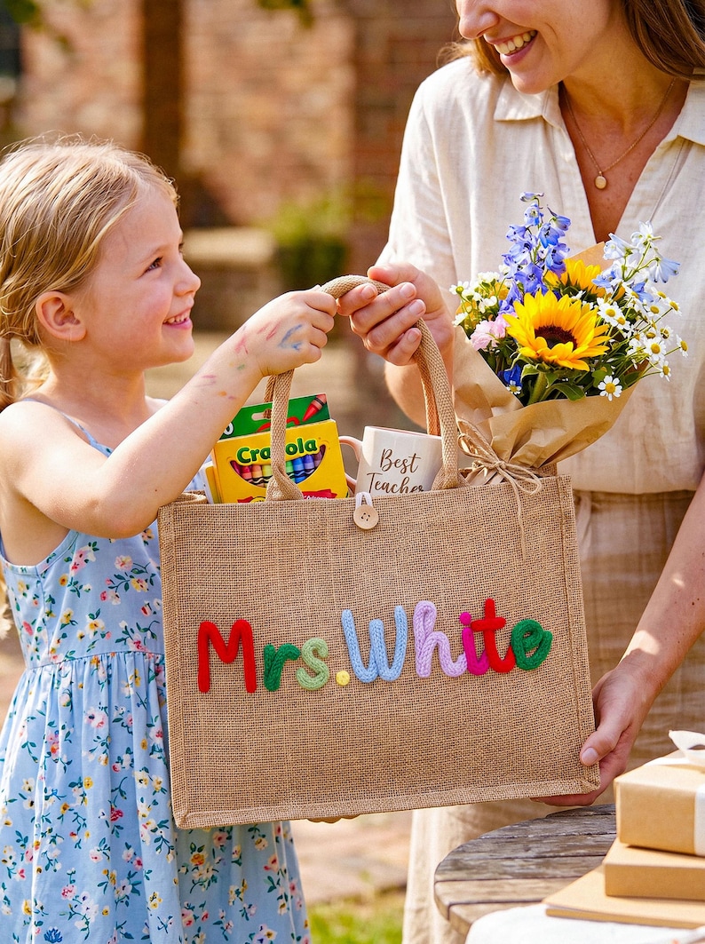 May include: A rectangular jute tote bag featuring the name Mrs. White in colorful, raised yarn lettering. The bag contains a box of Crayola crayons and a white mug with the text Best Teacher printed on it, alongside a bouquet of flowers.