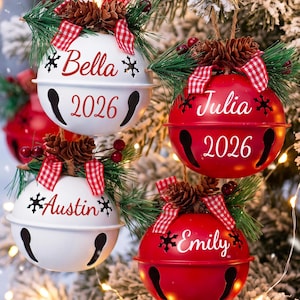 以下が含まれることがあります： Bella、Julia、Austin、Emilyの名前と2026年の年号が入った、赤と白の丸いベルのオーナメント4つ。各ベルの上部には、松の枝、小さな松ぼっくり、赤い実、赤と白のギンガムチェックのリボンが飾られています。