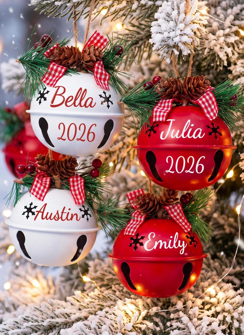 以下が含まれることがあります： Bella、Julia、Austin、Emilyの名前と2026年の年号が入った、赤と白の丸いベルのオーナメント4つ。各ベルの上部には、松の枝、小さな松ぼっくり、赤い実、赤と白のギンガムチェックのリボンが飾られています。