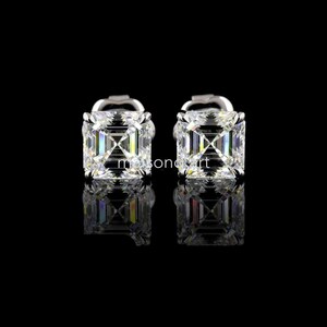 May include: A pair of square-cut, clear gemstone stud earrings. The earrings are set in a silver-colored metal and reflect light, creating a sparkling effect. The background is black, highlighting the earrings.