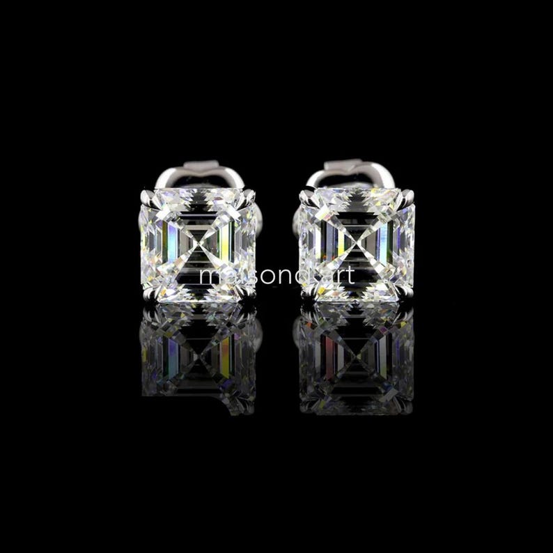 May include: A pair of square-cut, clear gemstone stud earrings. The earrings are set in a silver-colored metal and reflect light, creating a sparkling effect. The background is black, highlighting the earrings.