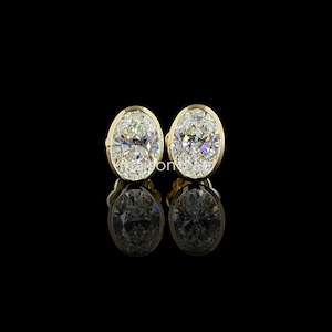 Könnte beinhalten: Ein Paar ovale Diamant-Ohrstecker in Gold gefasst. Die Diamanten sind klar und funkelnd und reflektieren das Licht. Die Ohrringe werden vor einem schwarzen Hintergrund präsentiert, mit ihren Reflexionen darunter.