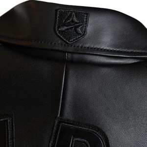 May include: Close-up of a black leather jacket. The jacket features a small, embroidered emblem on the collar and large, stitched letters on the lower portion. The leather appears smooth and the stitching is precise. The jacket is a classic piece of apparel.