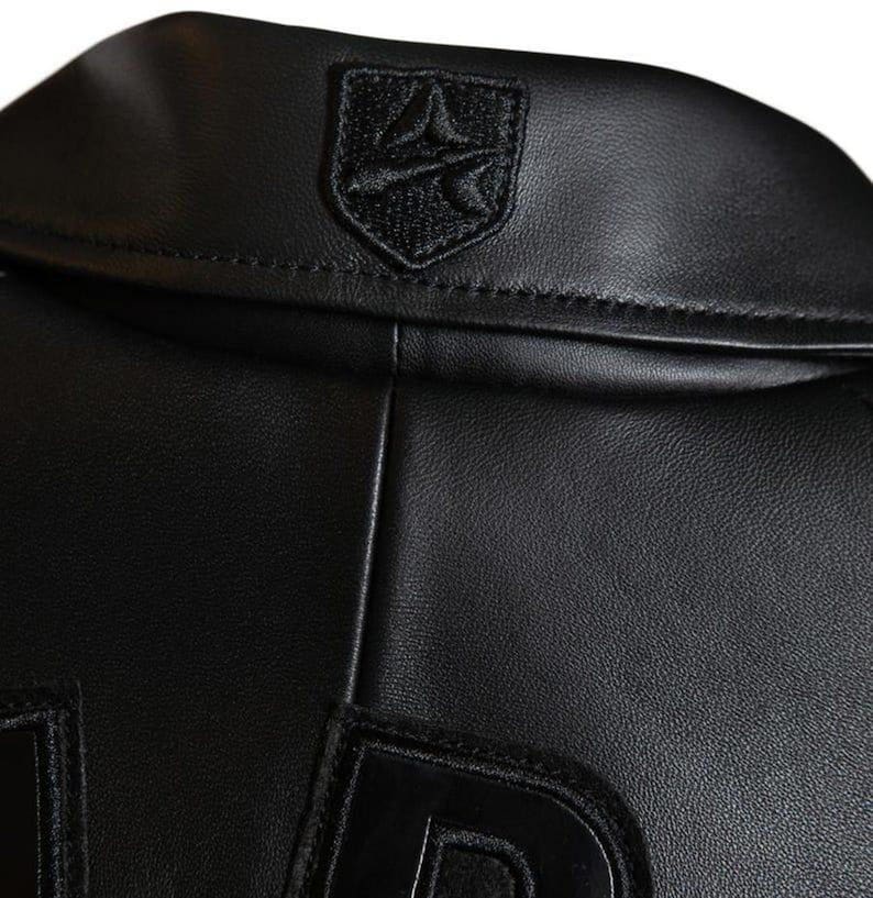 May include: Close-up of a black leather jacket. The jacket features a small, embroidered emblem on the collar and large, stitched letters on the lower portion. The leather appears smooth and the stitching is precise. The jacket is a classic piece of apparel.