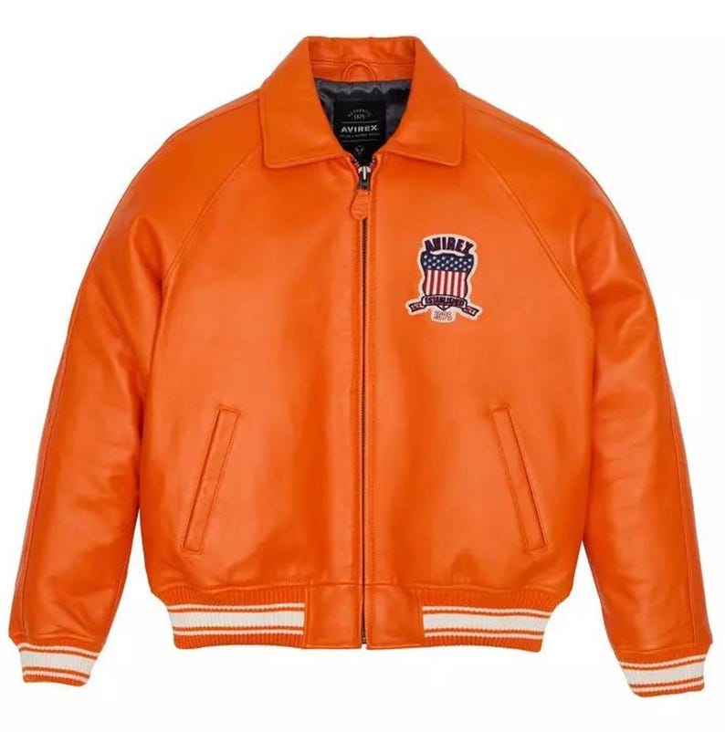 May include: A bright orange leather jacket featuring a front zipper closure, a classic collar, and ribbed cuffs and hem with white stripes. A patch on the left chest displays the text AVIREX with a shield graphic containing the American flag.