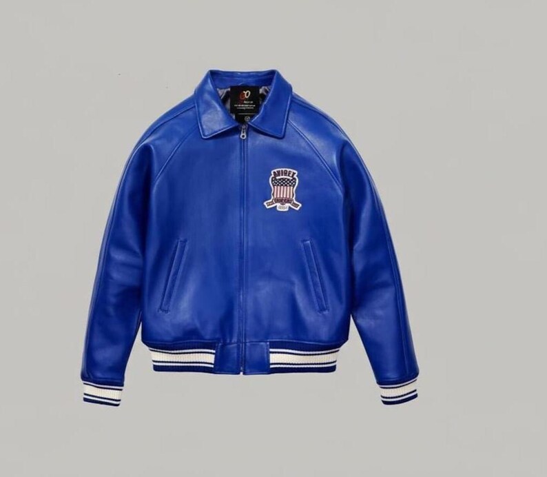 May include: A vibrant blue leather jacket featuring a classic collar, a full-length front zipper, and two side pockets. On the left chest, there is a patch displaying an American flag shield with the text AVIREX. The cuffs and hem have white stripes.