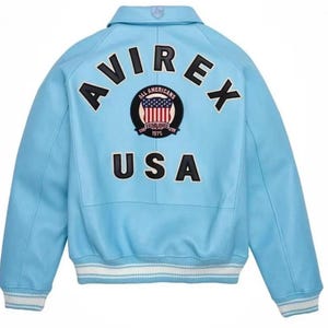 Puede incluir: Chaqueta de cuero azul claro con la palabra "AVIREX" en letras negras en la espalda. Un parche circular con "ALL AMERICANS" y "ESTABLISHED 1975" rodea un dise&ntilde;o de la bandera estadounidense. La palabra "USA" est&aacute; impresa debajo. Pu&ntilde;os a rayas blancas y azules.
