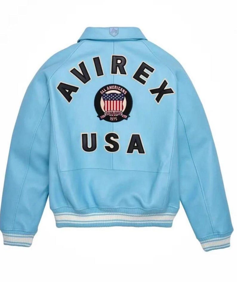 Puede incluir: Chaqueta de cuero azul claro con la palabra "AVIREX" en letras negras en la espalda. Un parche circular con "ALL AMERICANS" y "ESTABLISHED 1975" rodea un dise&ntilde;o de la bandera estadounidense. La palabra "USA" est&aacute; impresa debajo. Pu&ntilde;os a rayas blancas y azules.