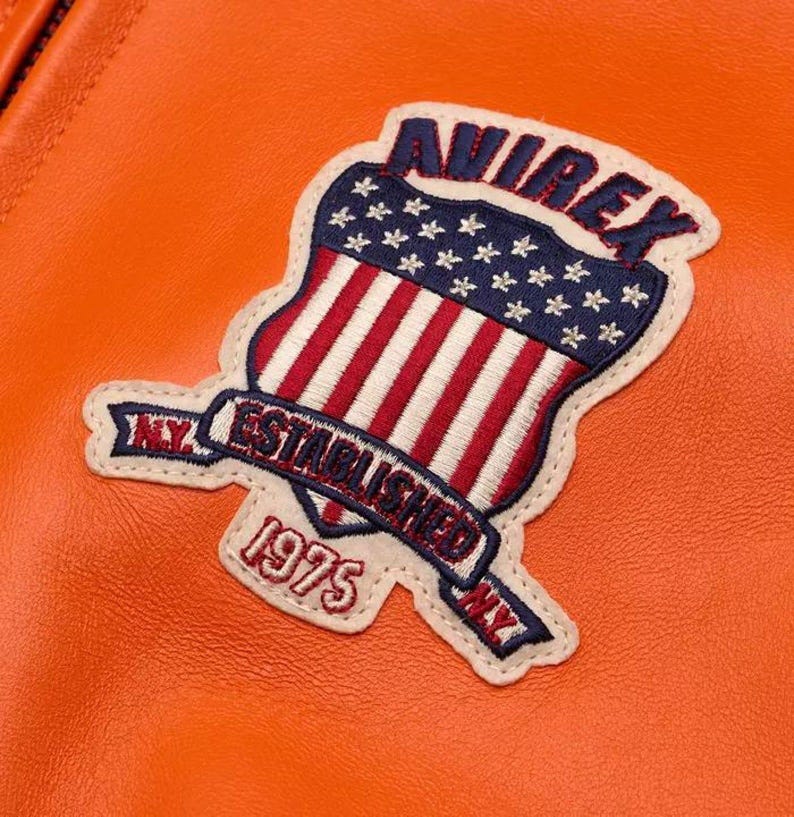 May include: A bright orange leather jacket featuring a large embroidered patch. The patch displays the text AVIREX, ESTABLISHED, 1975, and NY, alongside a shield graphic with red and white stripes and a blue field with white stars.