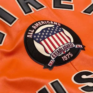 May include: A bright orange leather jacket featuring a large circular embroidered patch. The patch displays a shield with stars and stripes, the text ALL AMERICANS, ESTABLISHED 1975, and N.Y. Large black and white letters are visible on the jacket fabric.