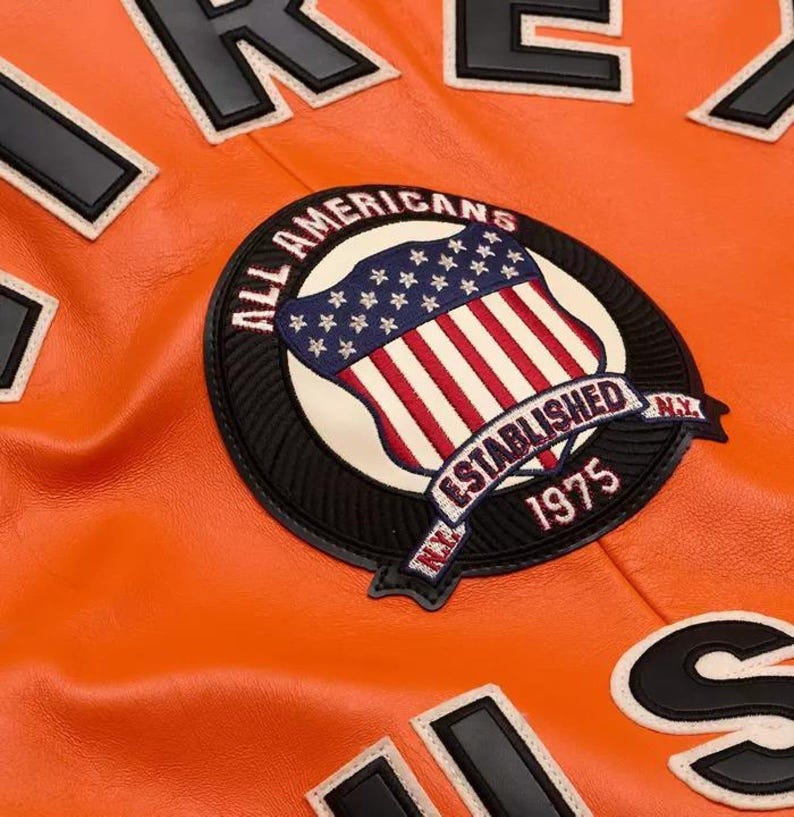 May include: A bright orange leather jacket featuring a large circular embroidered patch. The patch displays a shield with stars and stripes, the text ALL AMERICANS, ESTABLISHED 1975, and N.Y. Large black and white letters are visible on the jacket fabric.