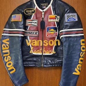 Può includere: Giacca da moto in pelle nera con diverse toppe, tra cui una bandiera americana, "Champion" e "Sunoco Race Fuels". La giacca presenta un pannello centrale rosso e marrone, strisce bianche e la scritta "Vanson" in giallo.
