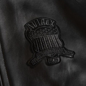 May include: Close-up of a black leather jacket with a textured surface. A black embroidered patch is sewn onto the jacket, featuring the word "AVIREX" above a shield design and the text "ESTABLISHED NY 1975".