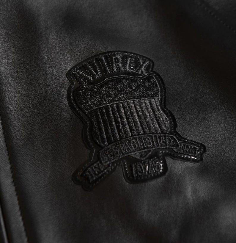 May include: Close-up of a black leather jacket with a textured surface. A black embroidered patch is sewn onto the jacket, featuring the word "AVIREX" above a shield design and the text "ESTABLISHED NY 1975".