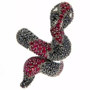 May include: A snake-shaped ring featuring a pavé setting of numerous small, round, faceted red and black stones. The design displays a coiled body with a distinct head, incorporating two small, clear, white stones as eyes set in a dark metal.
