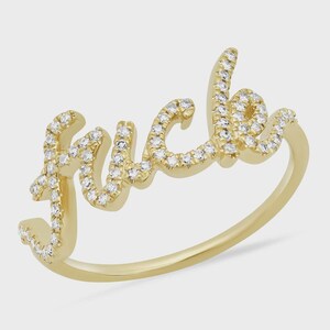 Puede incluir: Un anillo de tono dorado que presenta la palabra "fuck" escrita en una fuente cursiva. Toda la superficie de las letras está incrustada con pequeñas piedras claras y brillantes que captan la luz, sobre una banda de metal dorado pulido.