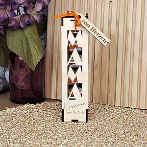 Puede incluir: Una caja de regalo de madera con la palabra "MAMA" recortada, llena de chocolate. La caja tiene una etiqueta que dice "von Herzen" y "Küsschen nur für Dich!" en escritura. Una cinta naranja está unida a la etiqueta. El fondo incluye un arco de madera y una estera tejida.