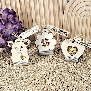 Puede incluir: Cajas de regalo de madera con forma de cerdo, flor y corazón, cada una con una pequeña etiqueta. El cerdo tiene el texto "Lass die Sau raus!". La flor tiene "Viel Glück". El corazón tiene "-lichen Glückwunsch".